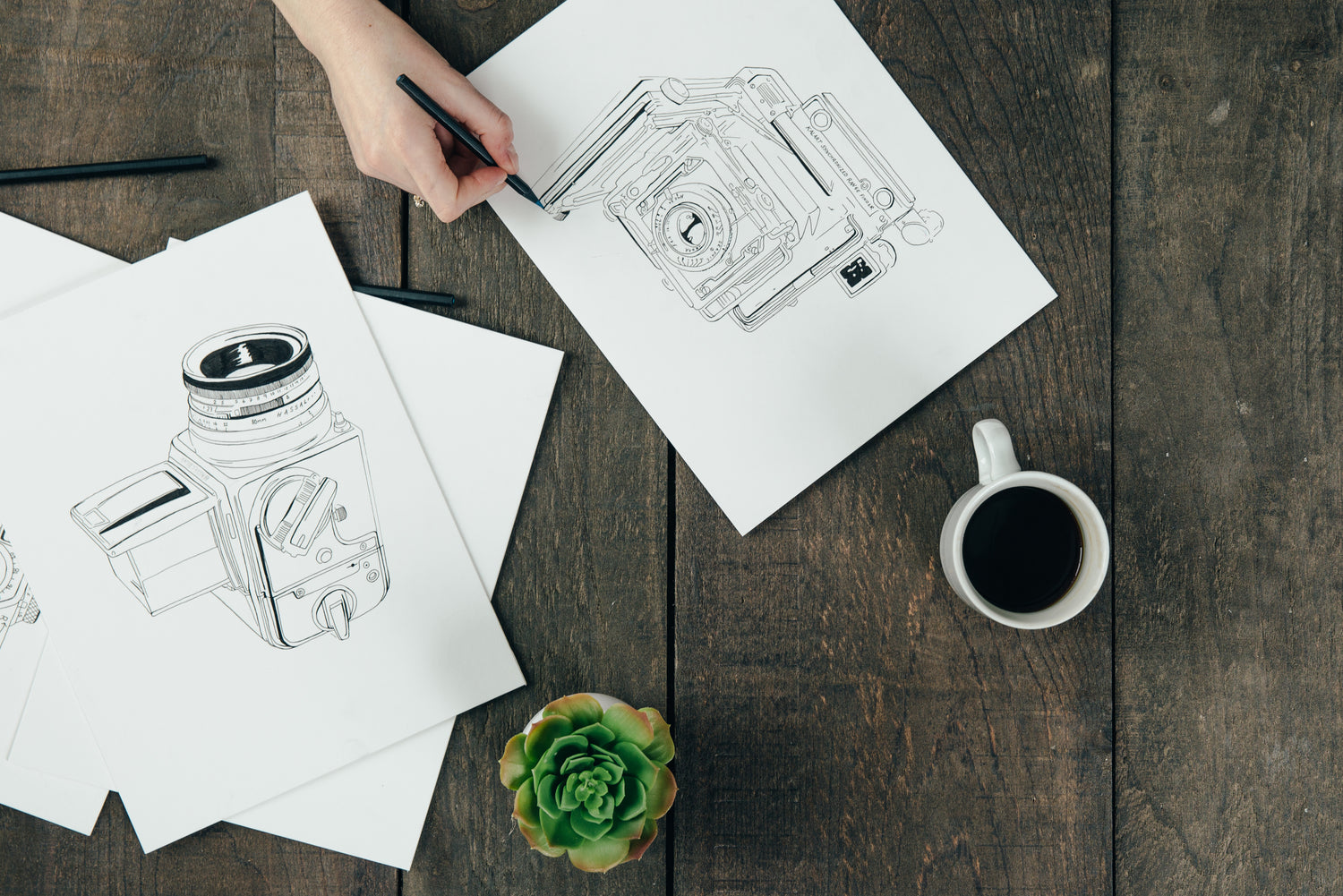 An image of illustrations drawn on paper of cameras with a cup of coffee on top of a wooden table